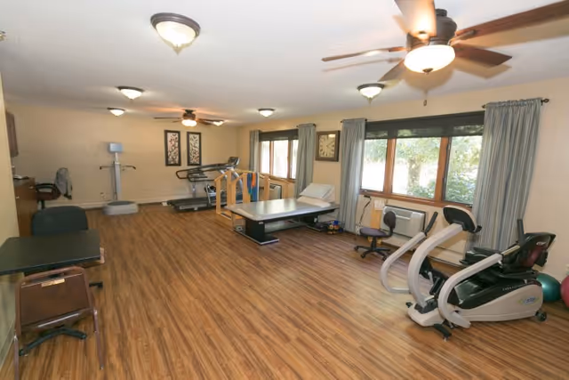 A spacious room with wooden flooring equipped with various exercise and rehabilitation equipment including a treadmill, stationary bike, therapy table, and parallel bars. The room has large windows with curtains, ceiling fans, and framed artwork on the walls.