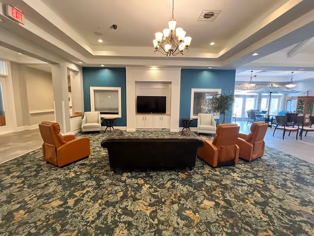 A spacious living room area with a patterned carpet, featuring a black sofa in the center, flanked by two brown armchairs on each side. Two white armchairs are placed against a blue accent wall with a flat-screen TV mounted in the middle. There are small side tables next to the white chairs and a chandelier hanging from the ceiling. The room opens into a dining area with tables and chairs visible in the background.