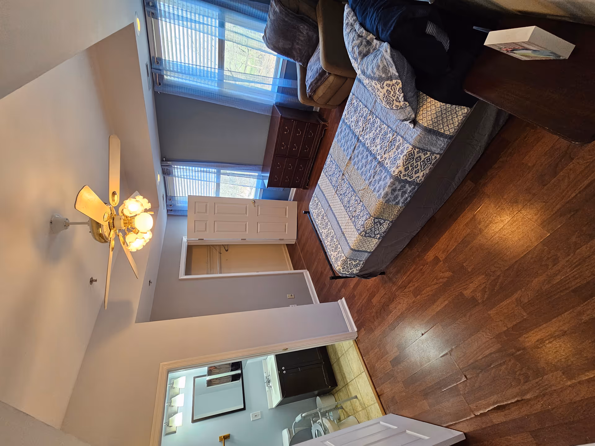 A bedroom in an assisted living facility with a single bed covered in a blue and white patterned quilt. The room has wooden flooring, a ceiling fan with lights, two windows with blue curtains, a wooden dresser, and a comfortable armchair. An open door reveals a small closet, and another door leads to a bathroom with a sink, mirror, and toilet.