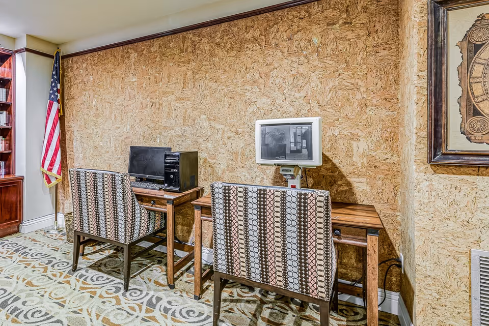 A small computer area in a senior living facility with two wooden desks, each with a computer and patterned chairs. The wall behind is covered with a textured cork-like material. An American flag stands in the corner next to a wooden bookshelf, and a framed artwork hangs on the right wall. The floor is carpeted with a decorative pattern.