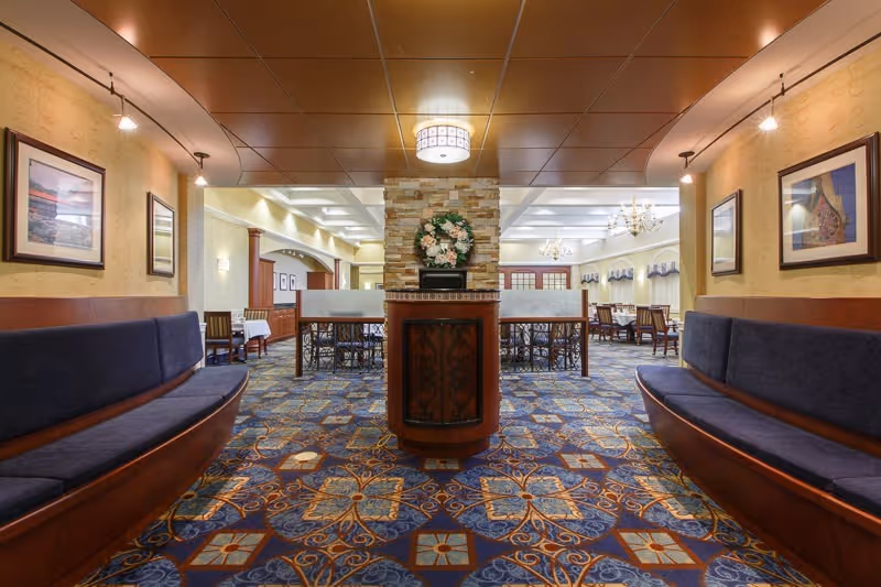 Well-lit communal dining/lounge area with curved bench seating, patterned carpet, a central decorative column with a wreath, and dining tables beyond.