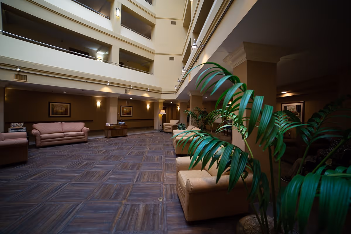Spacious indoor common area with multiple beige sofas and armchairs arranged along the walls and near large green plants. The room has a high ceiling with multiple floors visible, warm lighting, framed artwork on the walls, and a dark wood patterned floor.