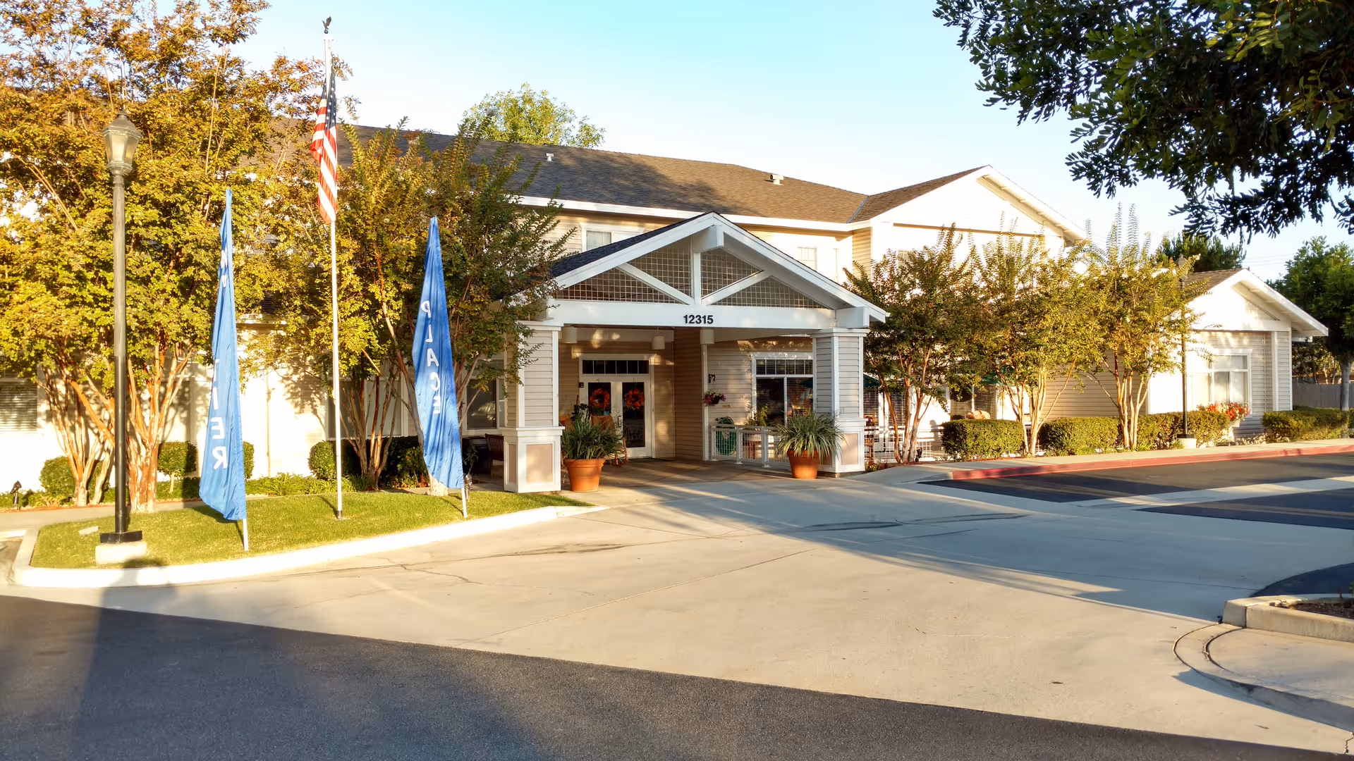 Front exterior view of a senior living facility named Discovery Commons Whittier, showing a covered entrance with the address number 12315 above it, surrounded by trees, two blue flags, and an American flag on flagpoles.