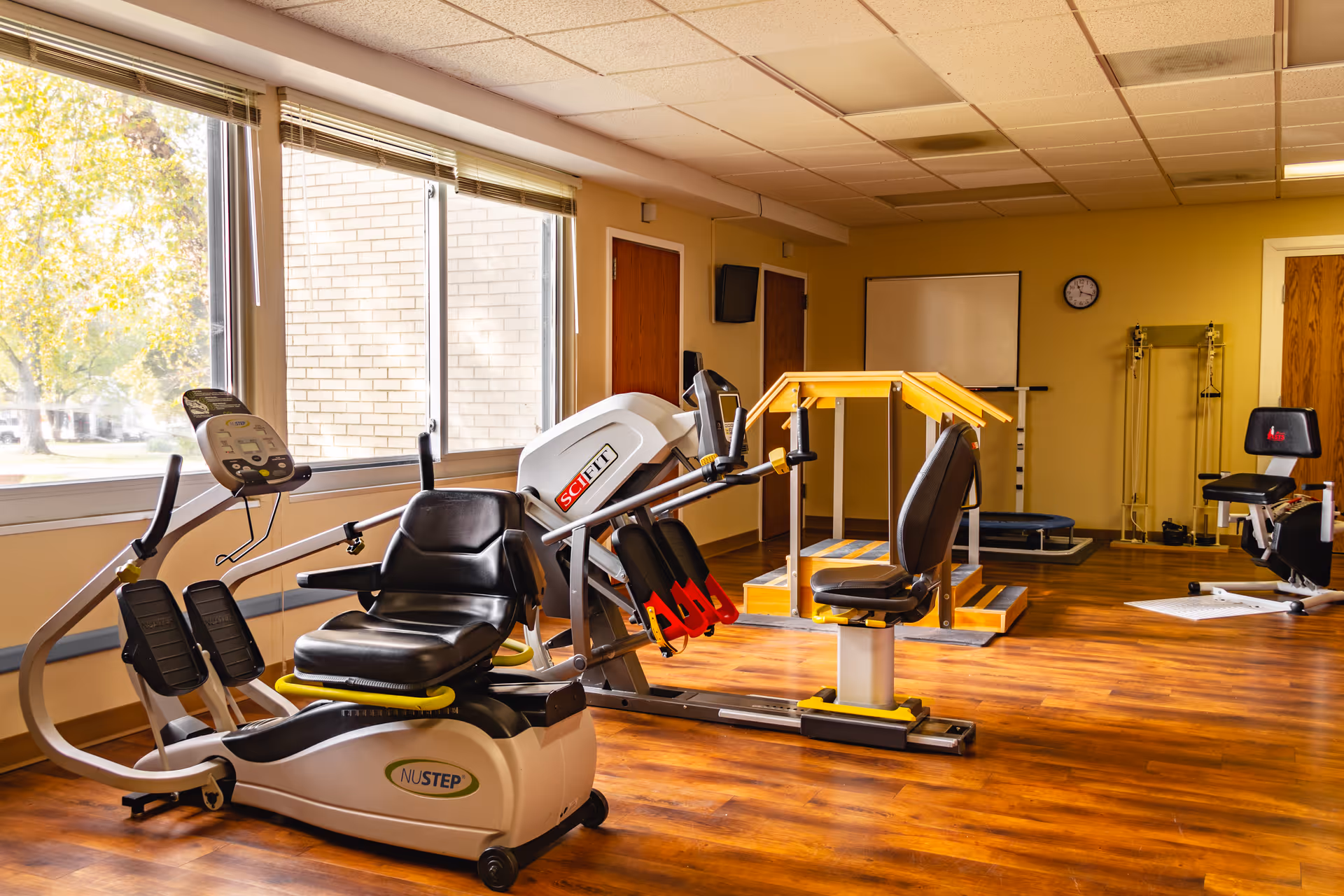 A well-lit exercise room with large windows showing trees outside. The room contains various fitness equipment including a NuStep recumbent cross trainer, a SciFit machine, a wooden balance or walking aid structure, and other exercise machines. The floor is wooden, and the walls are painted yellow with a clock and a whiteboard mounted on one wall.