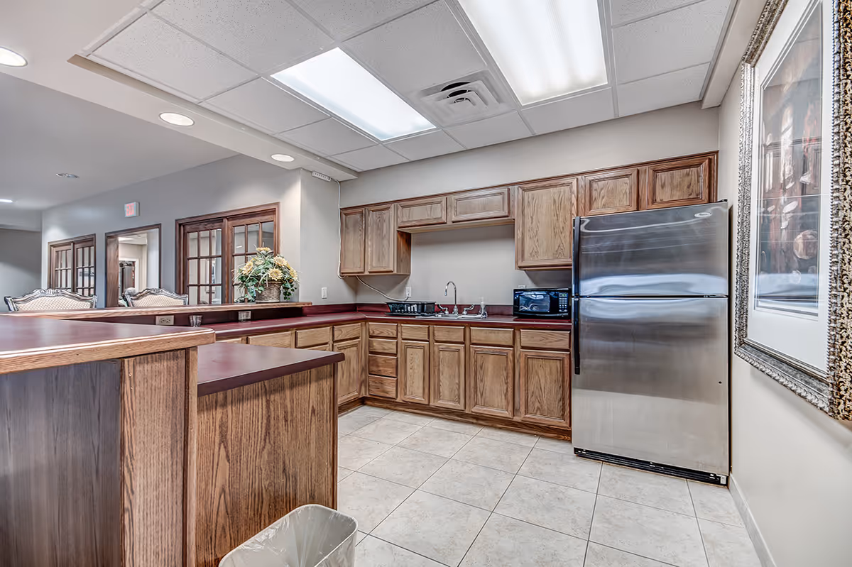 Interior view of a kitchen area with wooden cabinets, a stainless steel refrigerator, a microwave, a sink, and a countertop with a flower arrangement. The floor is tiled and there is a framed picture on the wall.