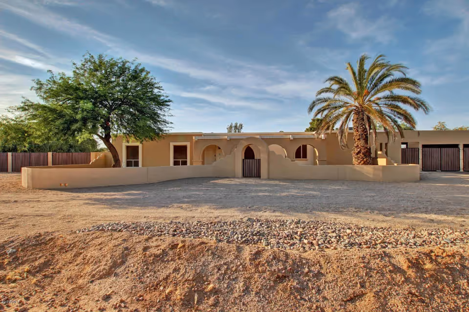 Single-story stucco building with arched entry and front courtyard wall, a large palm tree and desert landscaping under a blue sky.
