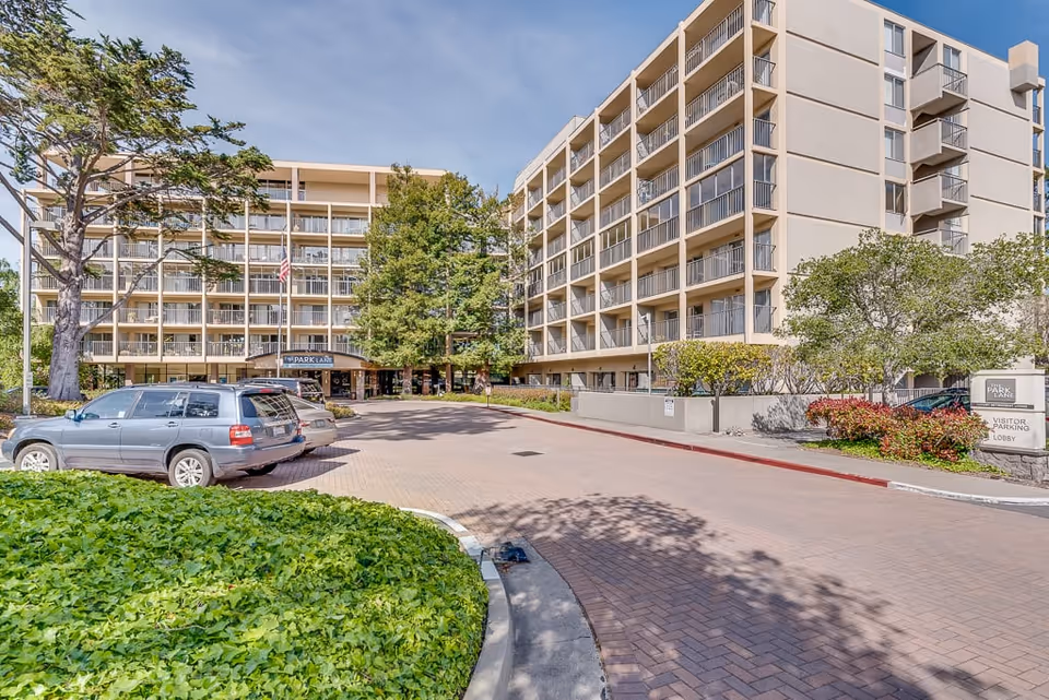 Exterior view of a multi-story senior living facility named Park Lane with balconies, surrounded by trees and greenery, with a driveway and parked cars in front under a clear blue sky.