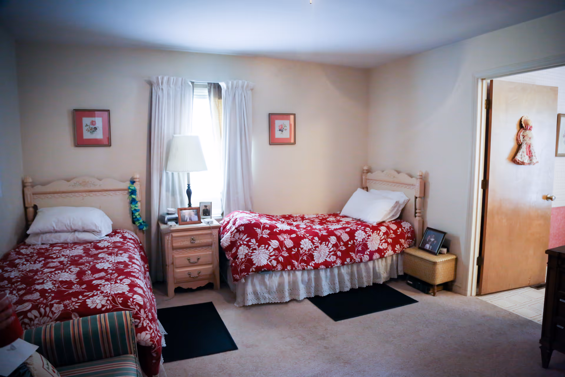 A bedroom with two single beds, each with a red and white floral bedspread. Between the beds is a wooden nightstand with a lamp, framed photos, and a phone. The room has beige walls, a window with white curtains, and two framed pictures above the beds. There is a door open to an adjoining room, and a striped armchair is partially visible in the foreground.