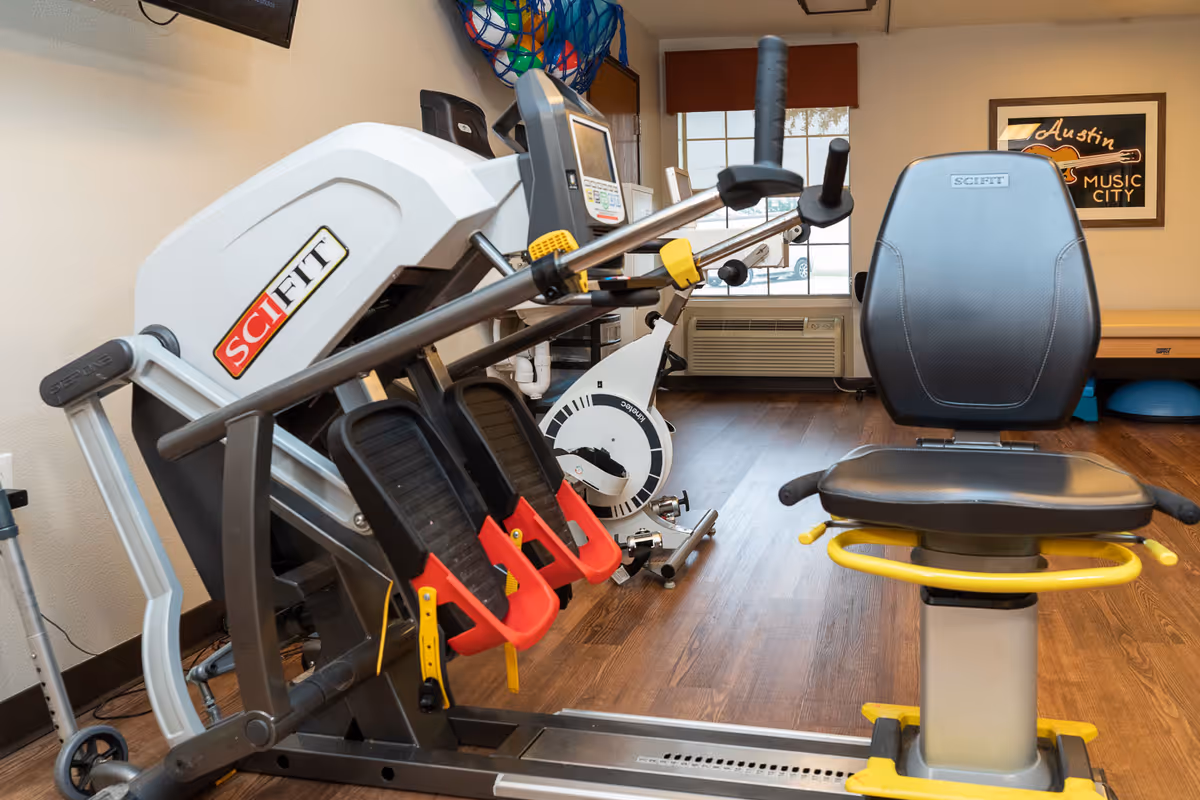 Exercise equipment including a SciFit rowing machine and a stationary bike in a room with wooden flooring, a window with blinds, an air conditioning unit, and a framed Austin Music City sign on the wall.