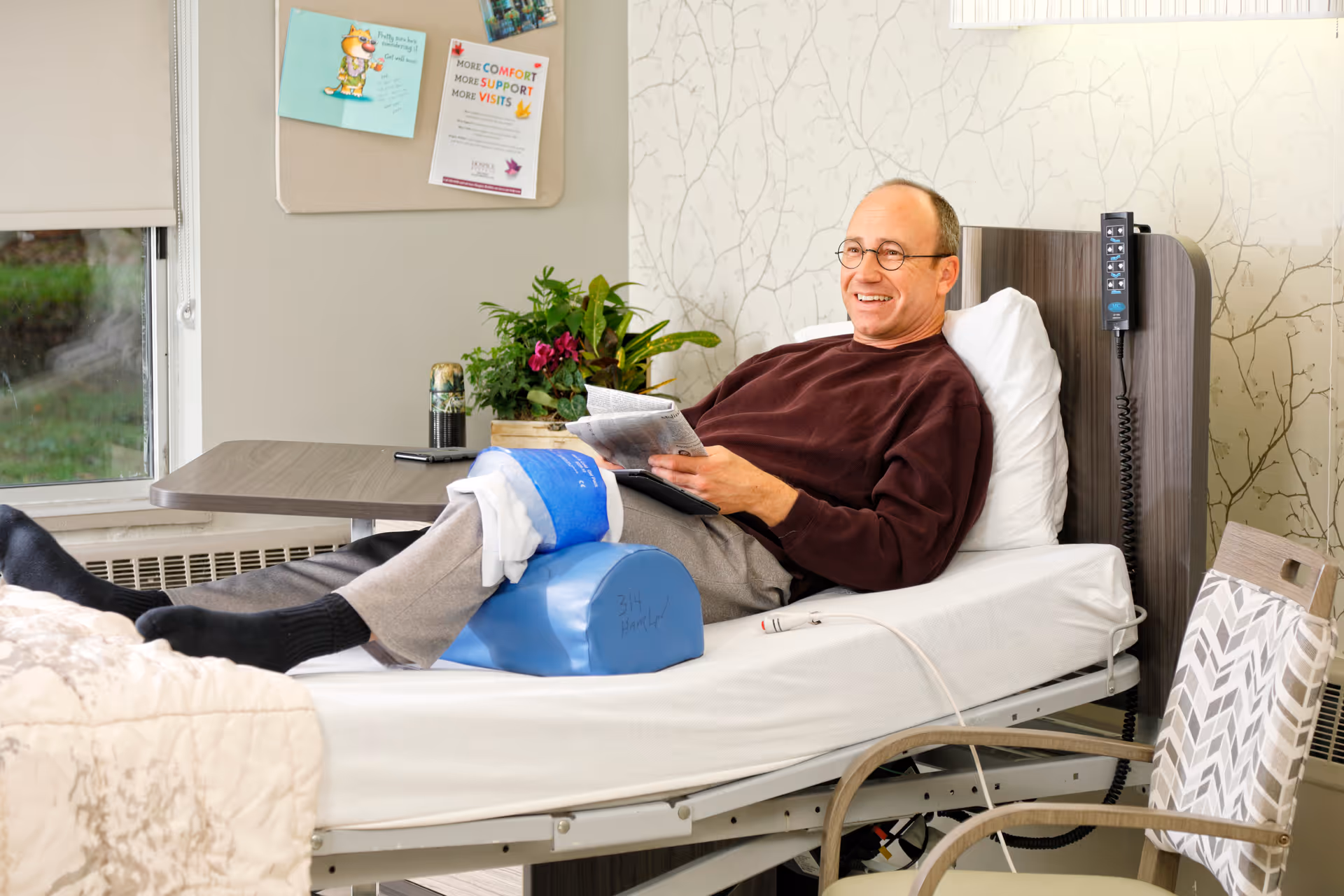 A man with glasses is lying in a hospital-style bed with his leg elevated on a blue cushion and wrapped in a white and blue bandage. He is smiling and reading a newspaper. The room has a window with a view of greenery outside, a small table with a plant and a drink, and a chair with a patterned cushion. The wall behind the bed has a subtle branch pattern wallpaper.