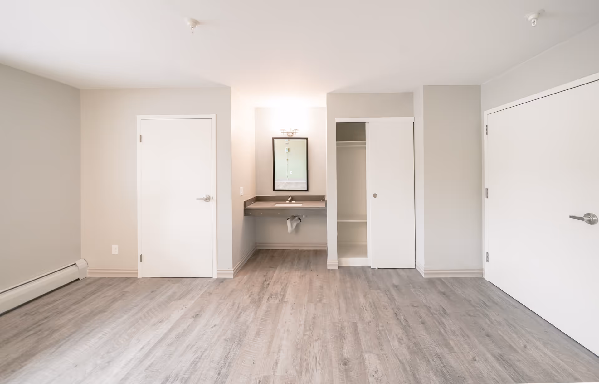 Empty room with light gray walls and wood flooring, featuring a small sink with a mirror above it in the center, a closed white door on the left, a closet with sliding doors on the right of the sink, and another closed white door on the far right.