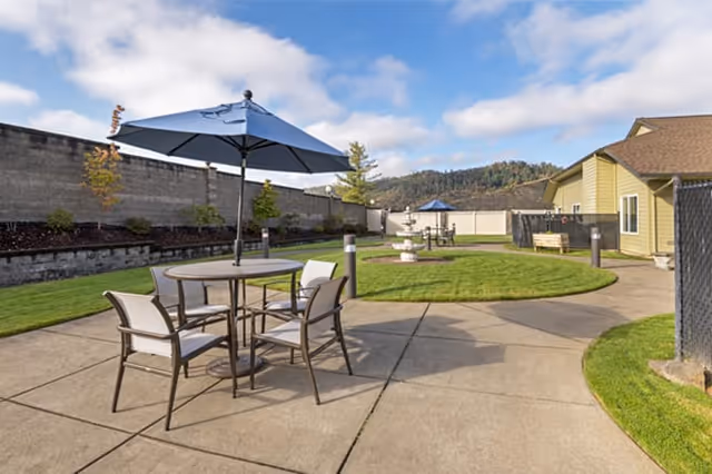 Outdoor patio area with round tables and chairs under blue umbrellas, surrounded by green grass and a concrete walkway. A building and a fence are visible in the background under a partly cloudy sky.