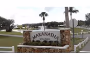 Stone-framed entrance sign reading MARANATHA VILLAGE, surrounded by lawn and palm trees.