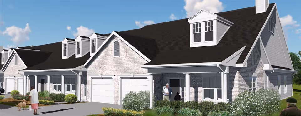 Exterior view of a residential-style senior living facility with multiple attached units featuring white brick walls, black shingled roofs, and white garage doors. There are bushes and greenery in front of the units, and a few people are visible walking near the building, including a person with a dog.