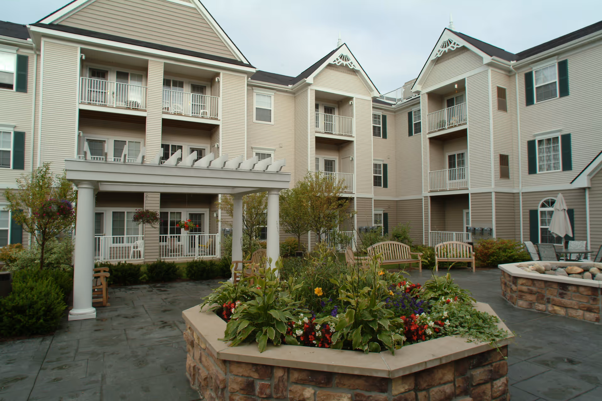 Outdoor courtyard area of a senior living facility with a stone planter filled with flowers and greenery in the center, surrounded by benches and chairs. The building has beige siding with white trim, multiple balconies, and green shutters on the windows.