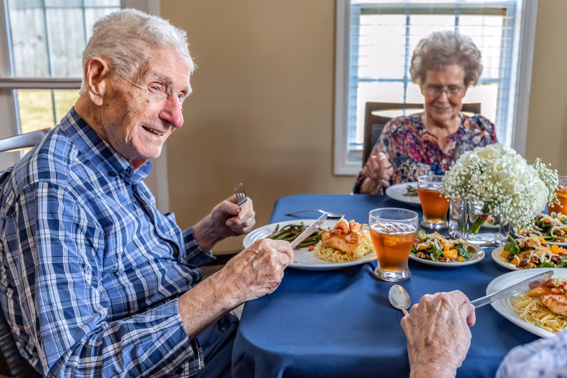 Two elderly individuals sitting at a dining table with plates of food including pasta, vegetables, and glasses of iced tea. The table is covered with a blue tablecloth and has a centerpiece of white flowers. They are in a well-lit room with windows in the background.