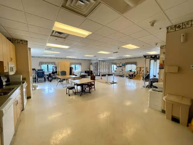 A spacious, well-lit rehabilitation or therapy room with exercise equipment, wheelchairs, tables, and chairs. The room has a tiled ceiling with fluorescent lights, large windows with valances, and a linoleum floor. There is a kitchenette area on the left side with cabinets, a microwave, and a dishwasher.