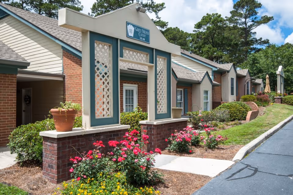 Exterior view of a residential building at Savannah Square with a sign reading 'Grey Moss Court 15-18'. The building has brick and beige siding with green trim, surrounded by landscaped bushes and blooming flowers under a partly cloudy sky.