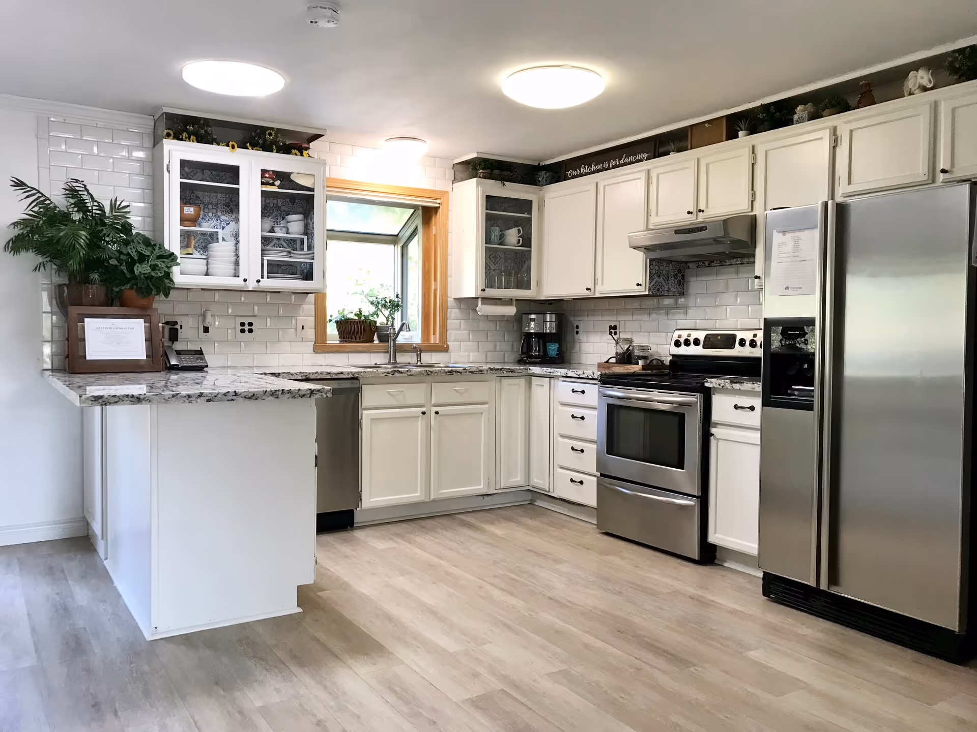 A bright and clean kitchen with white cabinets, stainless steel appliances including a refrigerator, oven, and dishwasher. There is a window above the sink with a wooden frame, and the countertops are granite. The kitchen has light wood flooring and white subway tile backsplash. There are some decorative plants and dishes visible in glass-front cabinets.