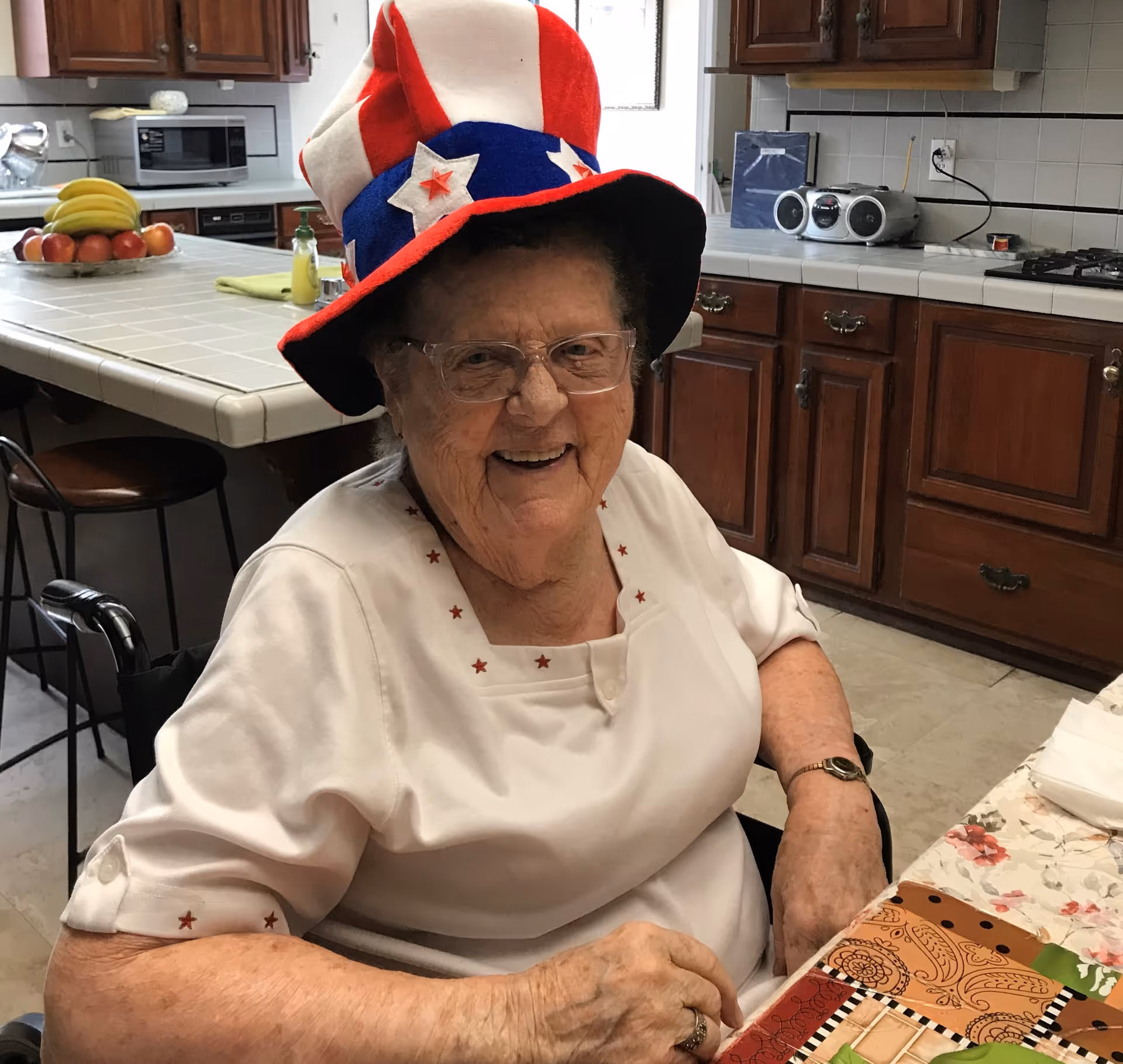 An elderly woman wearing glasses and a festive red, white, and blue hat with stars is smiling while sitting at a table in a kitchen. The kitchen has wooden cabinets, a microwave, a fruit bowl with bananas and apples, and a radio on the counter.
