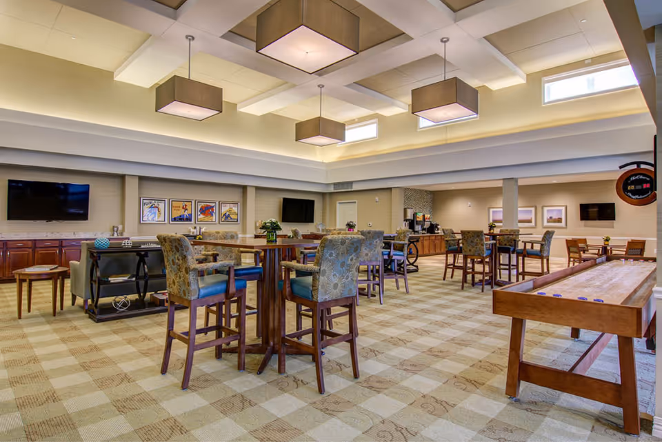 Spacious communal activity room with high ceilings, tables and chairs, TVs, and a shuffleboard table.
