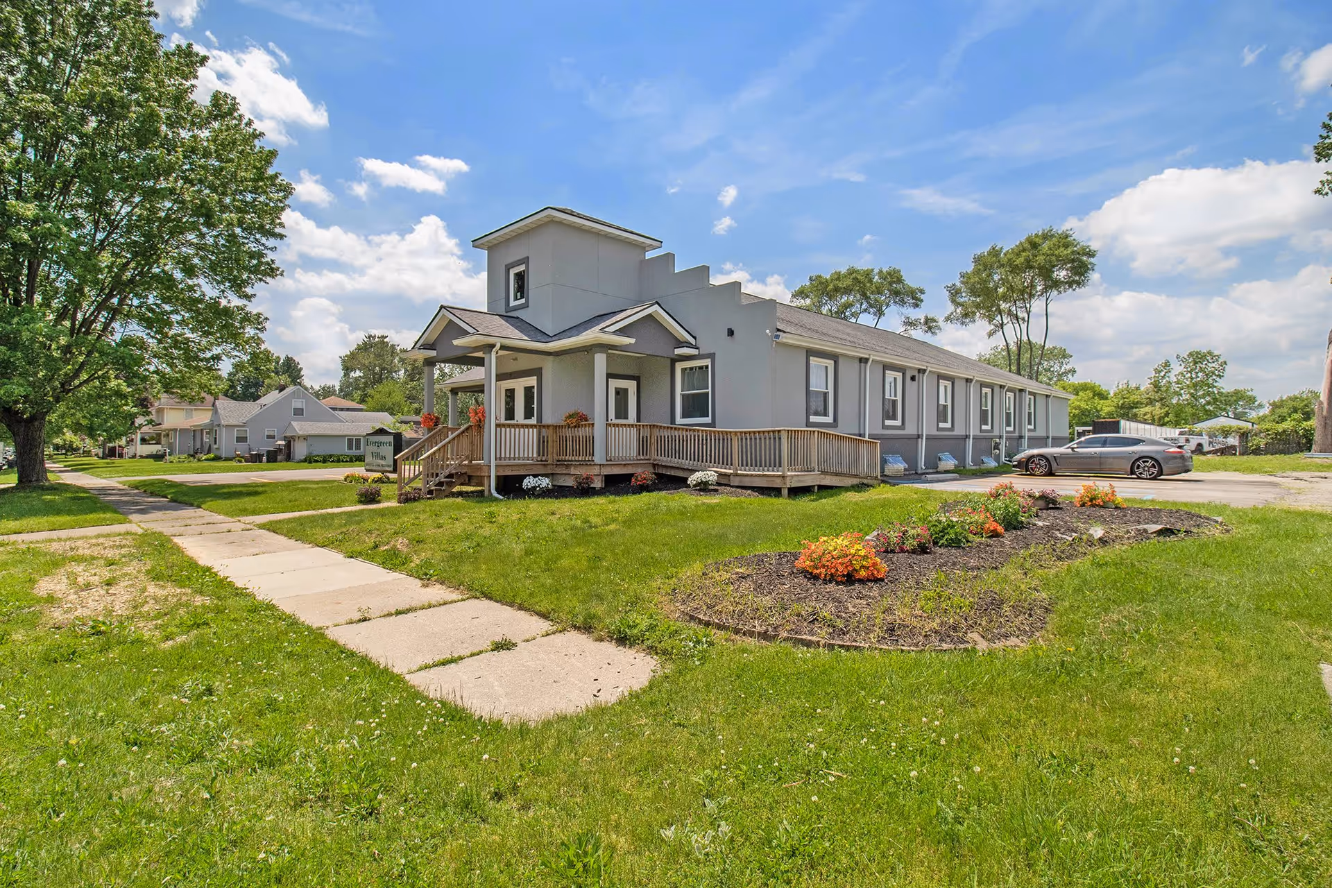 Front exterior of a single-story senior living building with a covered porch, ramp, lawn and flowerbeds under a blue sky.