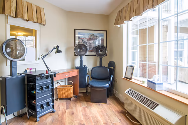 A small room with two black salon hair dryers, a black rolling cart with hair supplies, a wooden desk with a lamp, a wicker basket underneath, and a large window with beige curtains letting in natural light. The floor is wood, and there is a framed picture on the wall.