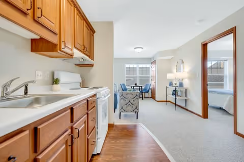 Bright furnished senior living apartment interior with a kitchenette in the foreground, a sitting area, and a bedroom visible through a doorway.