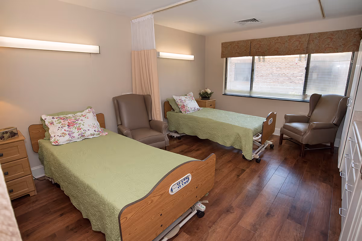 A shared healthcare bedroom with two hospital beds covered in green quilts, armchairs, bedside tables, and a large window.