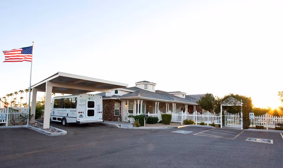 Exterior view of Arbor Rose Senior Care facility at sunset, showing a single-story brick building with a covered entrance where a white shuttle bus is parked. There is a white picket fence, an American flag on a flagpole, and a parking lot with handicap spaces in front.