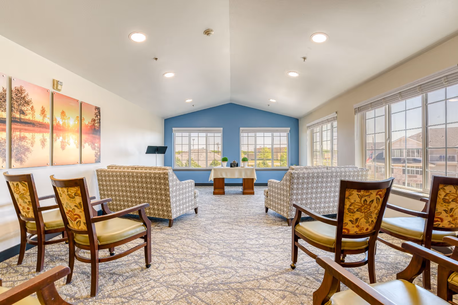 Bright communal lounge with sofas and wooden chairs arranged facing large windows and a table at the far end.