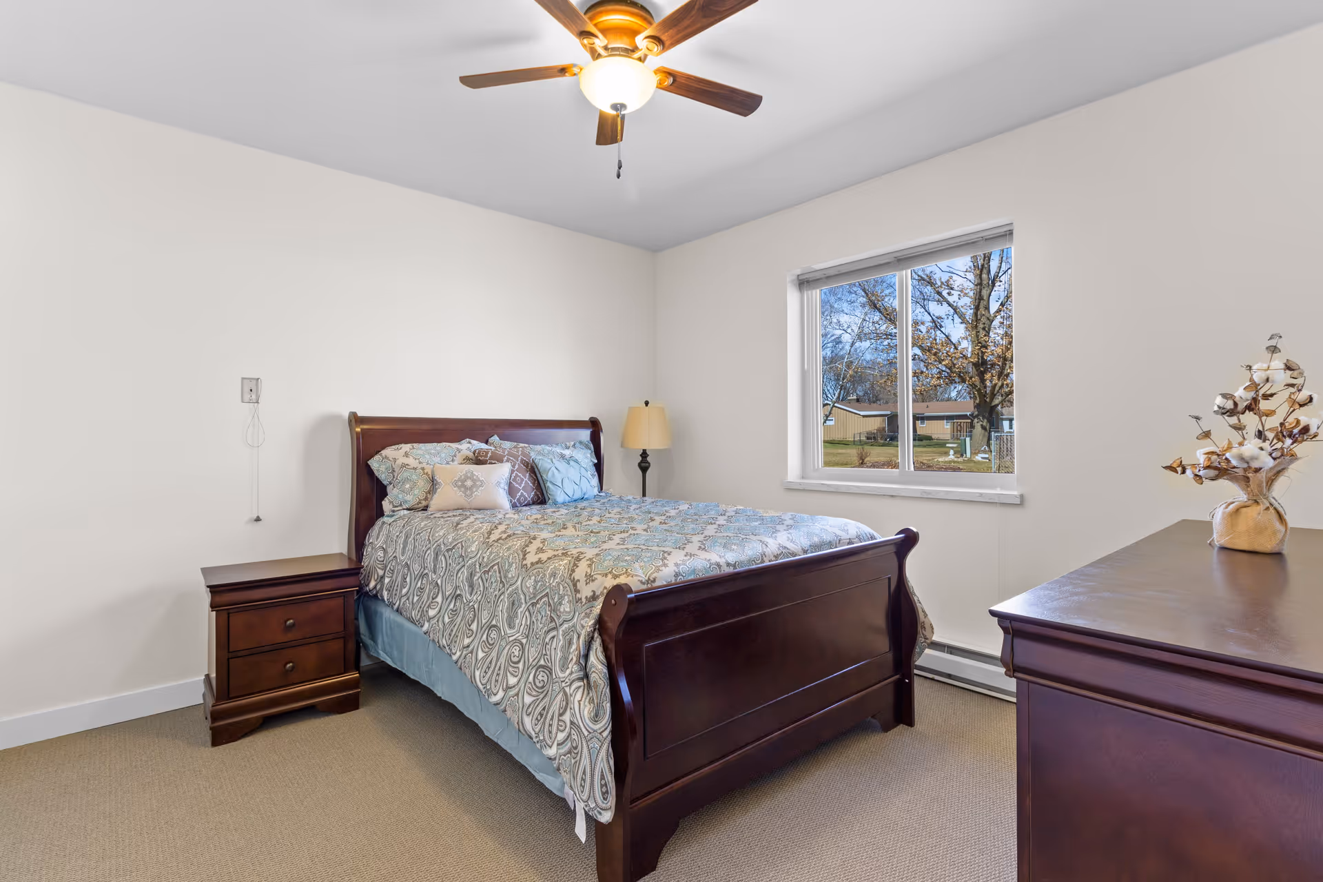 A bedroom with a wooden bed frame and a patterned bedspread in shades of blue and beige. There is a wooden nightstand with two drawers to the left of the bed and a floor lamp in the corner. A window on the right side shows an outdoor view with trees and buildings. A wooden dresser with a vase of cotton stems is visible in the foreground on the right side. The ceiling has a fan with a light fixture.