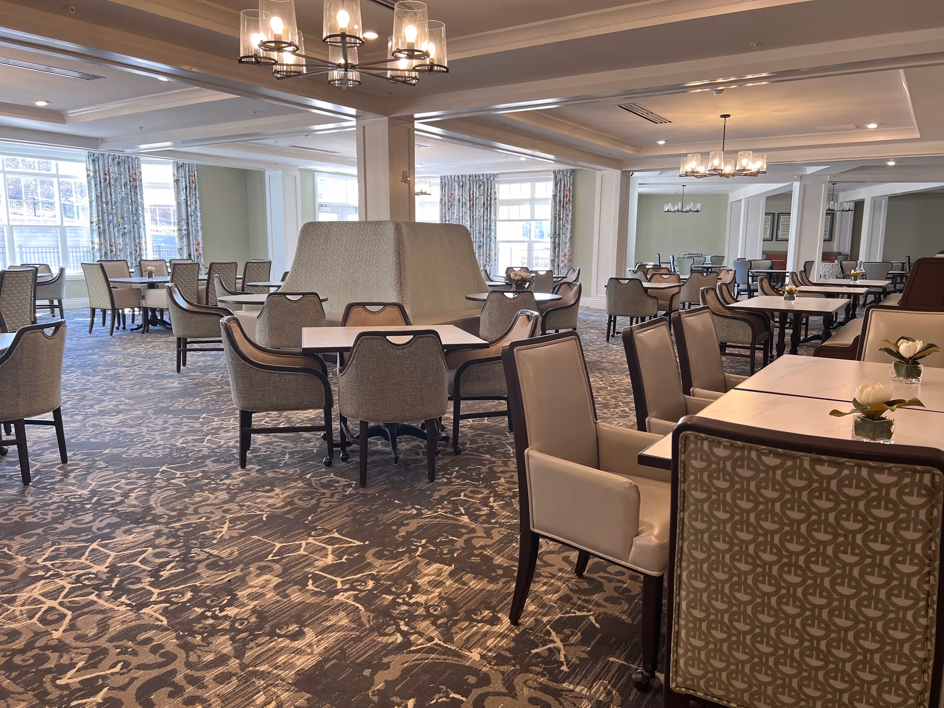 Large, well-lit dining room with multiple tables and upholstered chairs, chandeliers, patterned carpet, and windows with curtains.