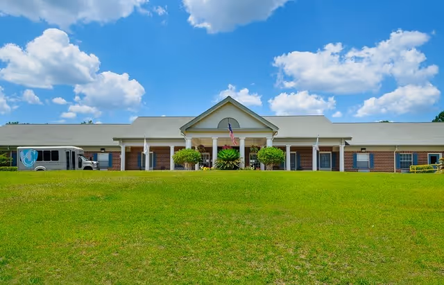 Front exterior view of a single-story brick building with a white roof and columns at the entrance, an American flag displayed above the entrance, a green lawn in the foreground, and a blue sky with scattered clouds.