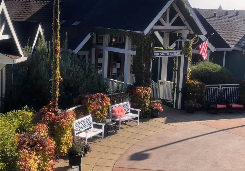 Outdoor seating area at Vista Terrace Of Belmont featuring white benches with red cushions, surrounded by lush greenery and climbing plants on a building with a dark roof. An American flag is displayed near the entrance under a white pergola with a sign that reads '100 SIXTH AVENUE'.