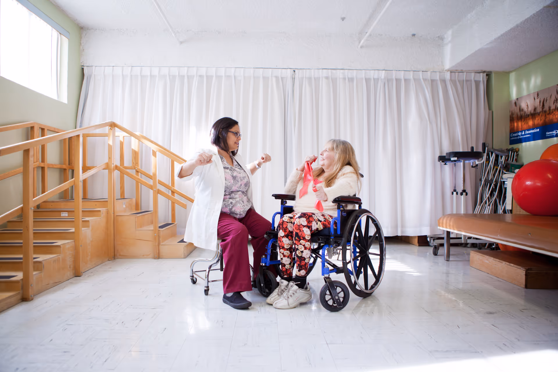 A healthcare professional in a white coat assists an elderly woman in a wheelchair with arm exercises using a red resistance band in a bright therapy room with wooden stairs and exercise equipment.