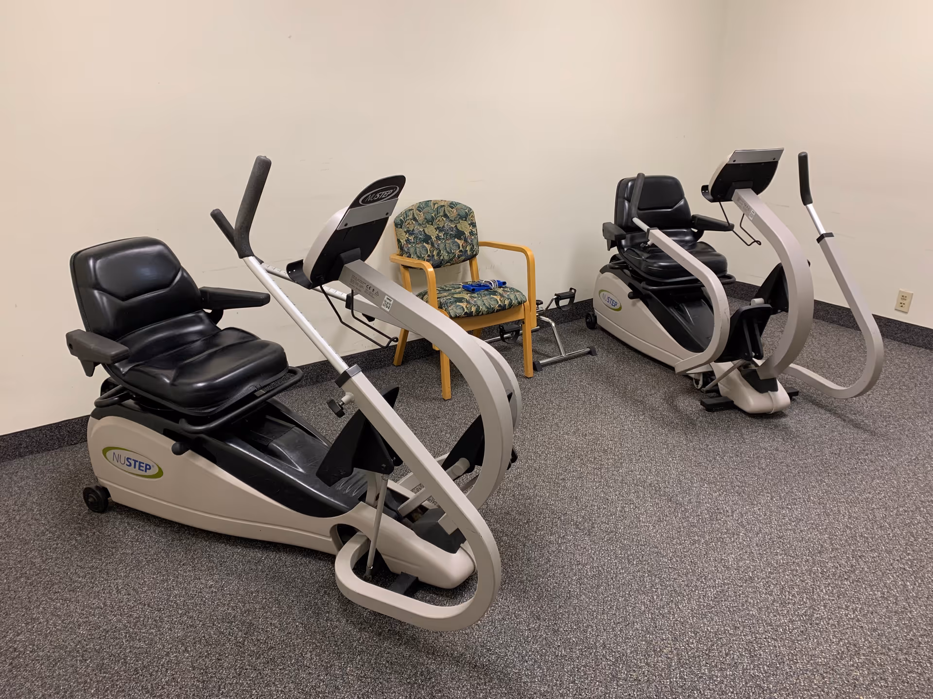 Small exercise room with two NuStep recumbent cross-trainers and a floral armchair against a beige wall.
