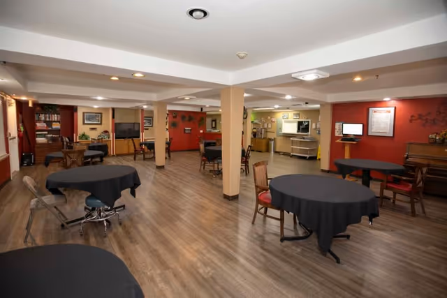 Interior view of a spacious common area with multiple round tables covered with black tablecloths and chairs around them. The room has wooden flooring, beige pillars, and red accent walls. There is a piano on the right side and a television mounted on the far wall. The area appears well-lit with ceiling lights and has a cozy, communal atmosphere.