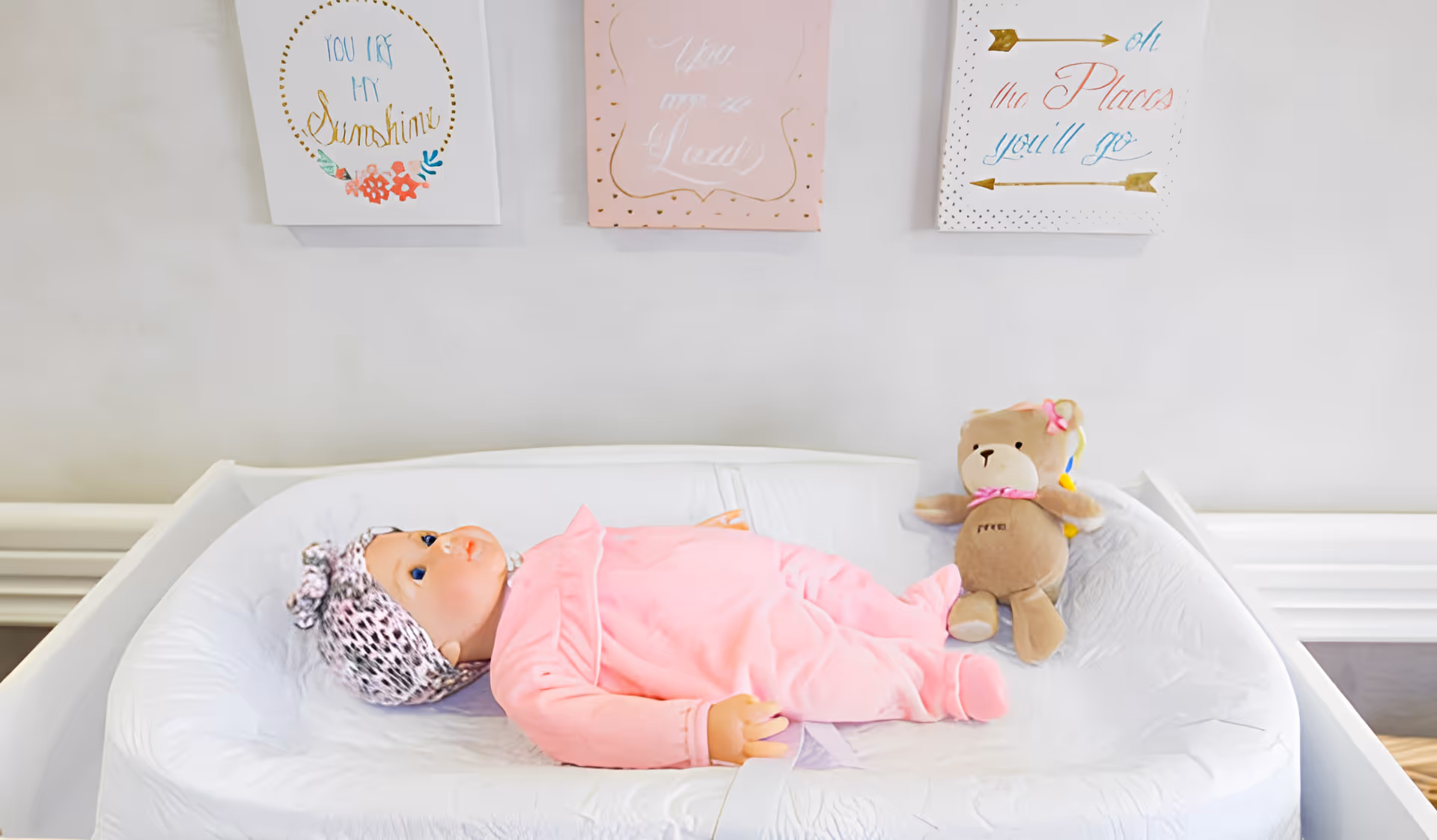 A baby doll dressed in pink lies on a white changing pad next to a small teddy bear beneath framed wall art.