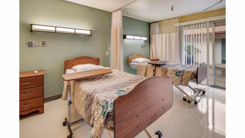 A healthcare facility room with two hospital beds separated by a privacy curtain. Each bed has a wooden headboard and footboard, a beige patterned blanket, and an overbed table. There is a wooden nightstand with drawers next to one bed. The room has green walls, overhead lighting, and a large window with vertical blinds allowing natural light to enter.