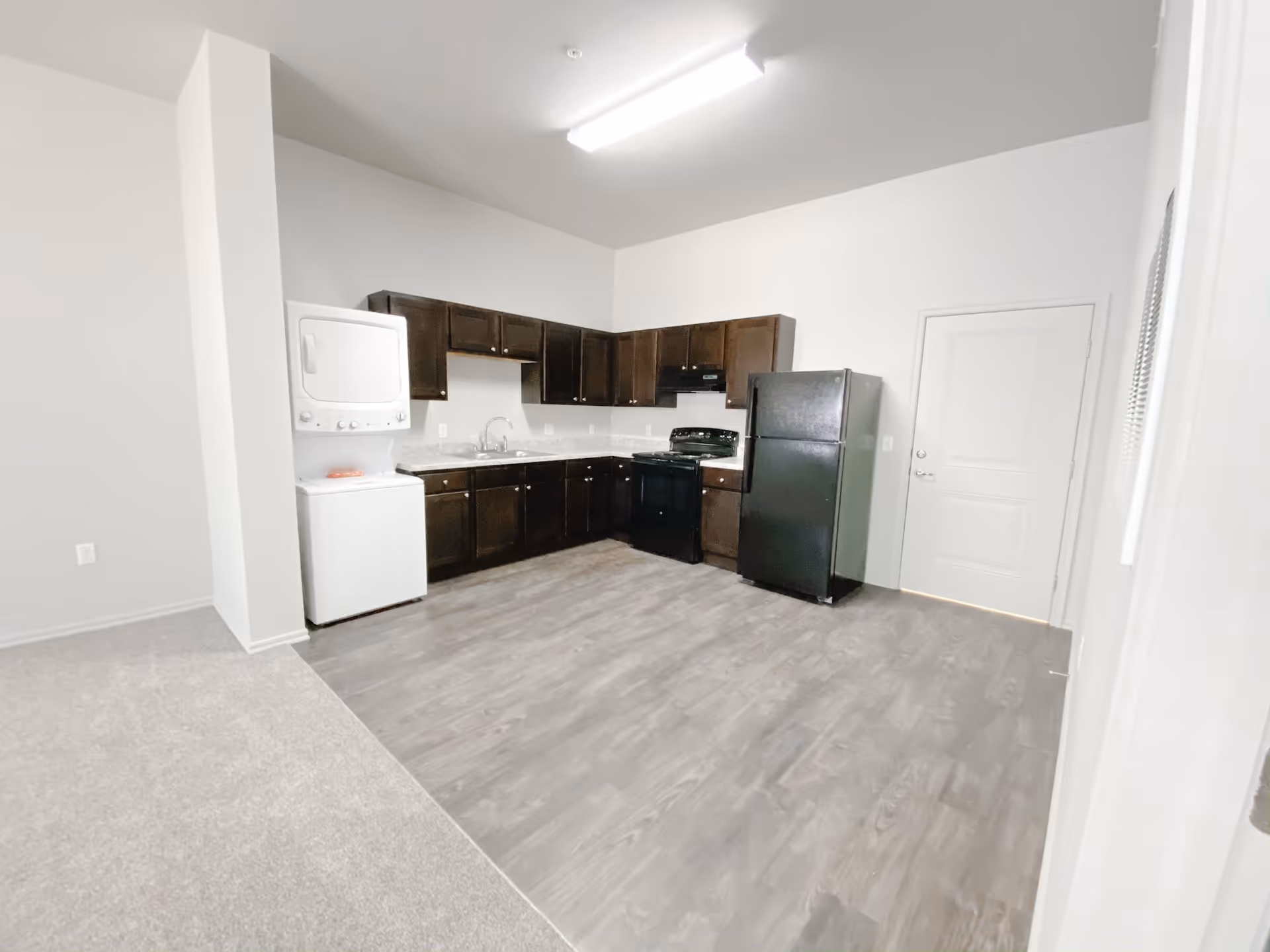 A modern kitchen area with dark brown cabinets, a black refrigerator, black stove, and a stacked white washer and dryer unit. The floor is a mix of light gray wood and carpet, and the walls are painted white. There is a closed white door to the right of the refrigerator.