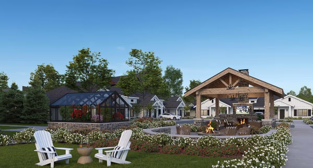 Outdoor seating area at The Inn at University Village Assisted Living Community featuring white Adirondack chairs with striped cushions, a small round table, a fire pit surrounded by chairs, a covered pavilion with a fireplace and TV, a greenhouse, and landscaped gardens with flowers and trees under a clear blue sky.