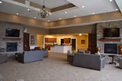 Spacious senior living community lounge with sofas, two stone fireplaces and wall-mounted TVs, a chandelier, and an open reception/kitchen area in the background.