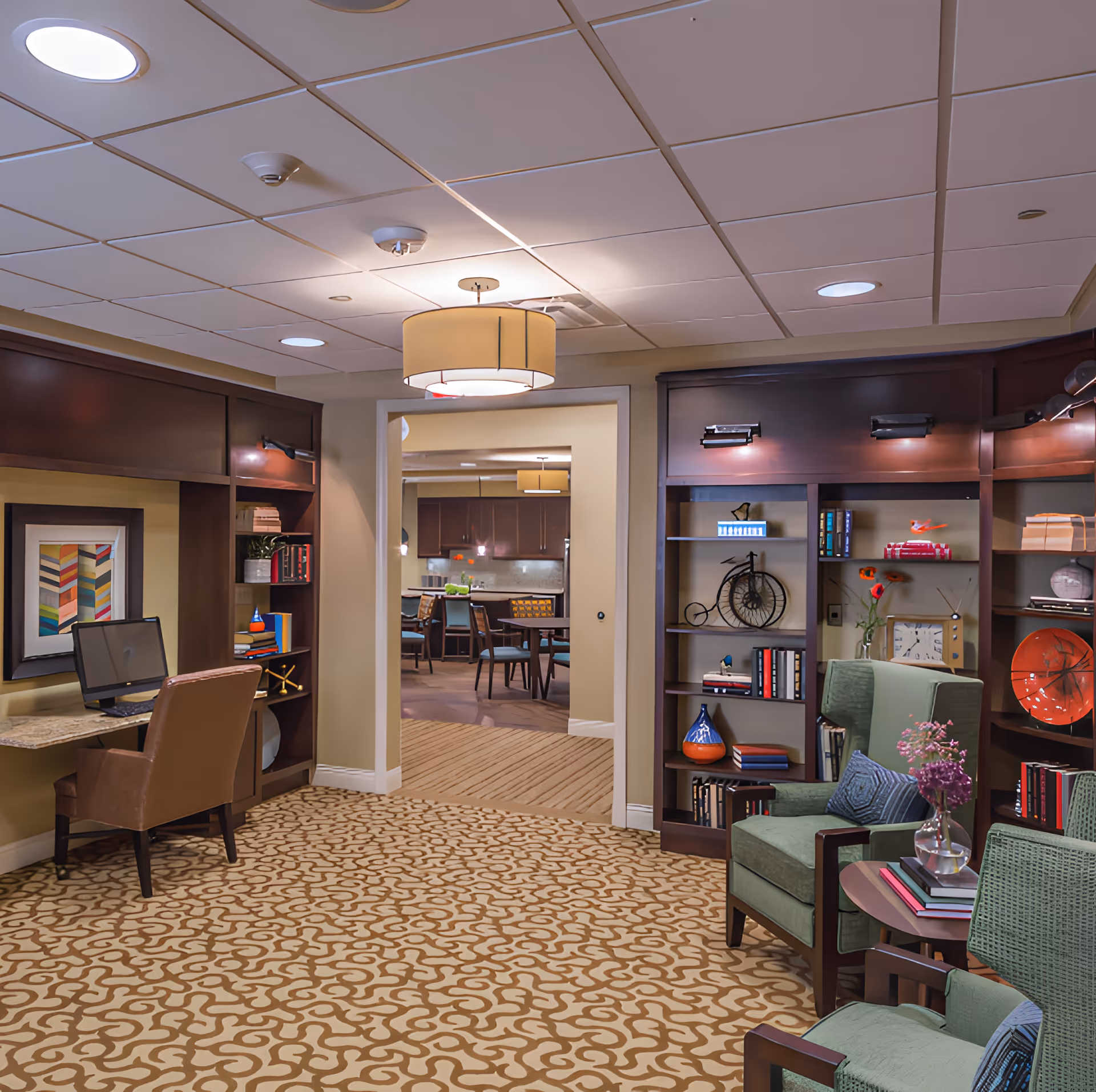 A cozy interior common area with patterned carpet, two green armchairs with cushions, a small round table with books and a vase of flowers, and built-in dark wood bookshelves filled with books and decorative items. A desk with a computer and a brown chair is on the left side. In the background, there is an open doorway leading to a dining area with tables and chairs and a kitchen with dark cabinetry.