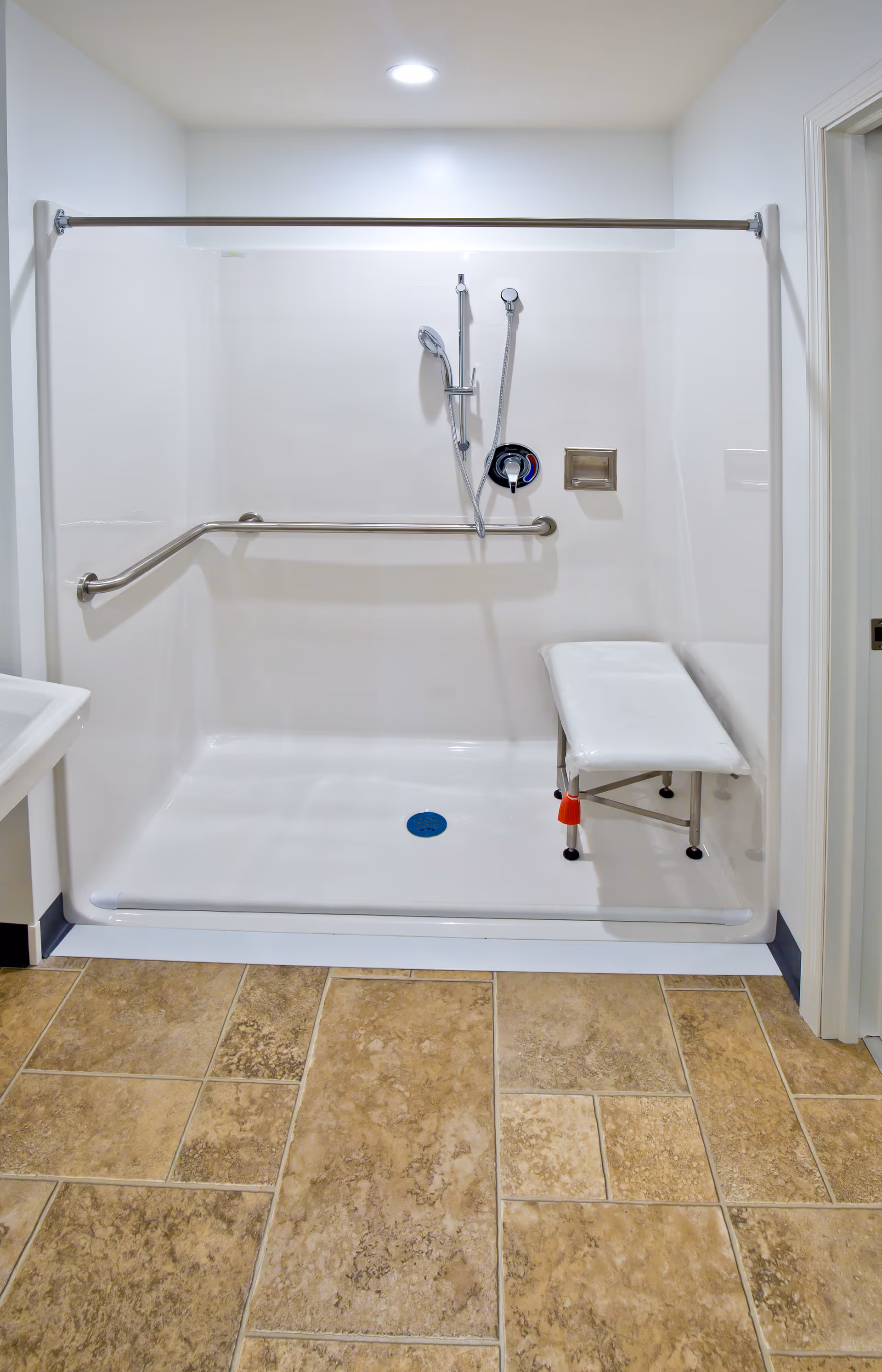 Accessible shower area with a white foldable shower bench, a handheld showerhead, a grab bar along the back wall, and a tiled floor outside the shower.