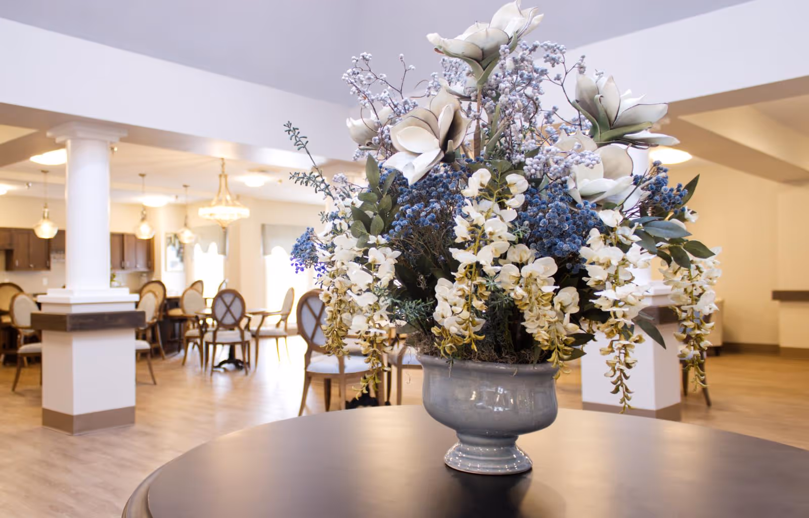 A large floral arrangement with white and blue flowers in a gray vase placed on a round table in the foreground. In the background, there is a spacious dining area with wooden chairs and tables, white columns, and warm lighting fixtures including chandeliers and pendant lights.
