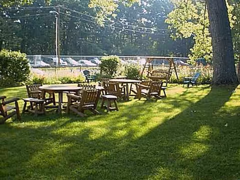 Sunlit outdoor lawn with wooden tables, chairs, and a swing set near trees.