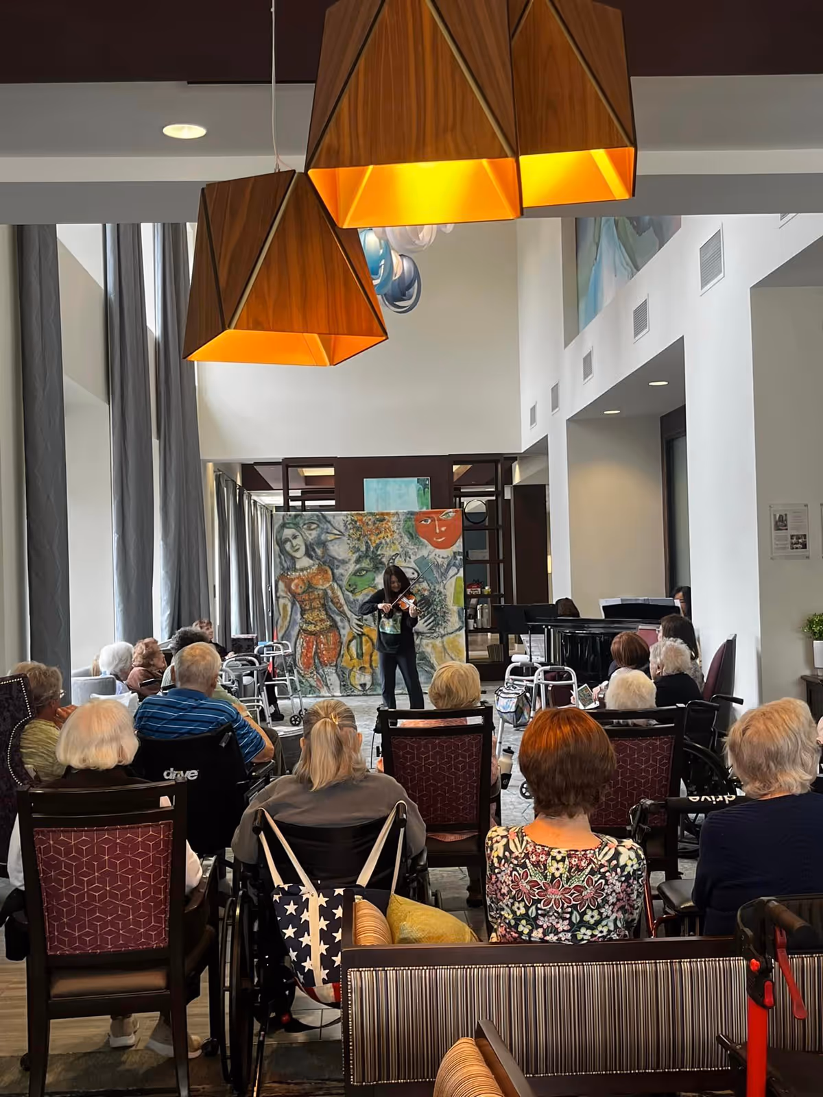 A group of elderly people seated in chairs and wheelchairs watching a woman playing the violin in a spacious, well-lit room with large windows and modern hanging lights. There is colorful artwork on the wall behind the performer.