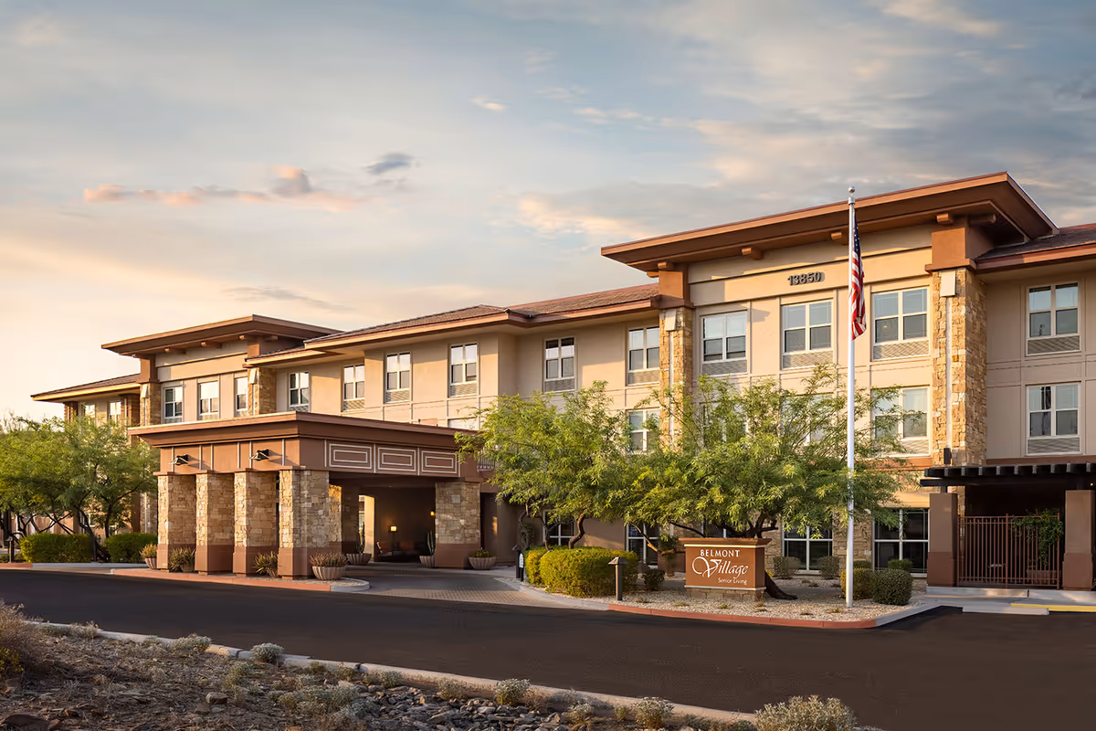 Front exterior of the Belmont Village Senior Living building in Scottsdale showing a three-story facade with a covered entrance, stone columns, flagpole, and landscaping.