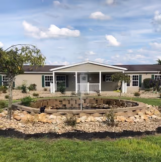 Single-story beige building with a covered entrance and white railings, surrounded by a landscaped garden featuring a circular stone water fountain in the foreground under a partly cloudy sky.